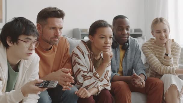 Medium-long of African, Asian, Mixed-Race and Caucasian friends sitting on couch at home, carefully watching captivating TV program, smiling and talking - Footage, Video