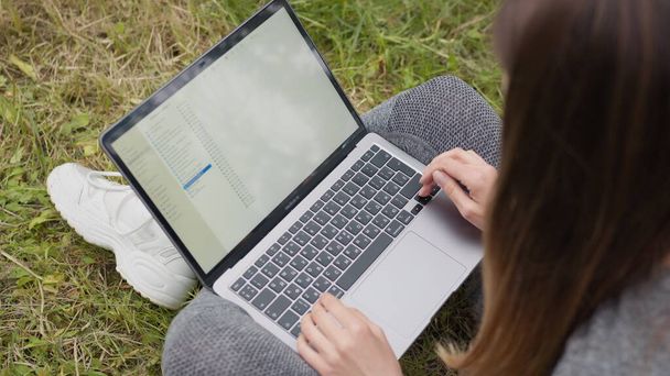 Top view Dziewczyna pracująca na laptopie na ulicy, siedząca na trawie. Dziewczyna Student lub Freelancer przygotowuje prace, sortuje pliki na komputerze, pracuje zdalnie z domu i biura. Ręce na klawiaturze - Zdjęcie, obraz