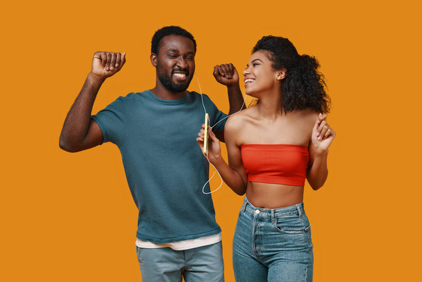 Beautiful young African couple smiling and listening music while standing against yellow background - Foto, Imagem