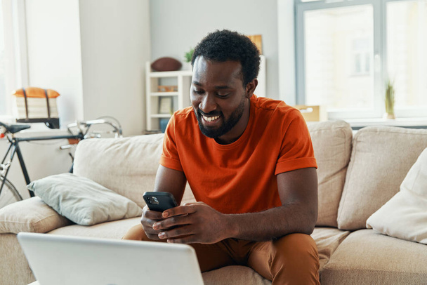 Carefree young African man using smart phone and smiling while spending time at home - Foto, Imagen