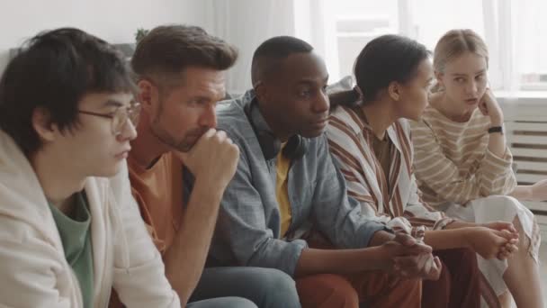 Średni długi z pięciu młodych, wielonarodowych ludzi siedzących na kanapie w salonie w domu, oglądających niewidzialną telewizję, mieszaną rasową kobietę rozmawiającą z białym przyjacielem - Materiał filmowy, wideo