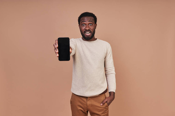 Handsome young African man in casual clothing showing his smart phone and looking surprised - Φωτογραφία, εικόνα