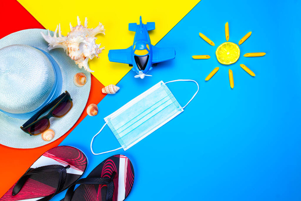 Medical mask and beach accessories with sunglasses, passports, straw hat, seashell flip flops and toy plane on colored background. Travel and tourism during the covid pandemic. Summer vacation. - Photo, Image