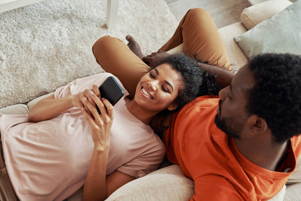 Top view of young African woman using smart phone and smiling while spending time at home with her boyfriend - Fotó, kép