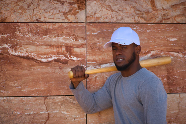 Afro-Américain en casquette blanche avec batte de baseball reposant sur son épaule sur un fond de pierre. - Photo, image
