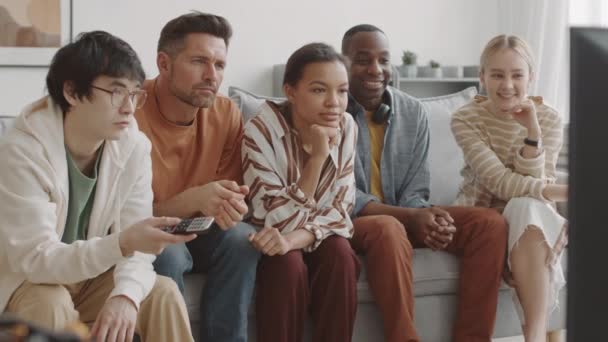 Middellange van Afrikaanse, Aziatische, gemengde-ras en blanke vrouwen en mannen zitten op de bank in de woonkamer, zorgvuldig kijken naar boeiende tv-programma, glimlachen en praten - Video