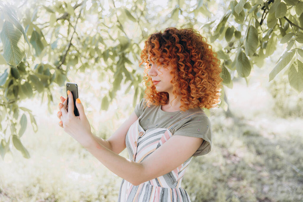 Redhead κορίτσι με afro μπούκλες με ένα τηλέφωνο σε ένα πράσινο πάρκο κάνει μια selfie και χαμόγελα, κάνει μια παρουσίαση smartphone - Φωτογραφία, εικόνα