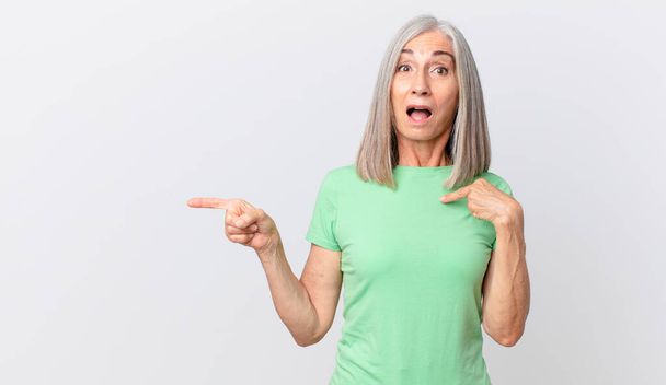 middle age white hair woman looking shocked and surprised with mouth wide open, pointing to self and pointing to the side - Photo, image