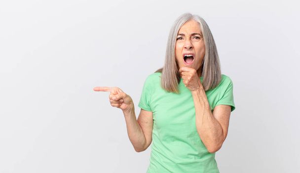 middle age white hair woman with mouth and eyes wide open and hand on chin and pointing to the side - Foto, Imagem
