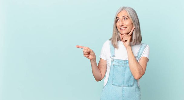 middle age white hair woman smiling happily and daydreaming or doubting and pointing to the side - Фото, изображение