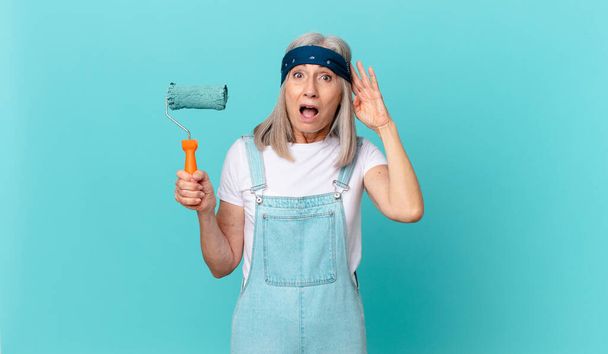 middle age white hair woman looking happy, astonished and surprised with a roller painting a wall - Foto, Imagem