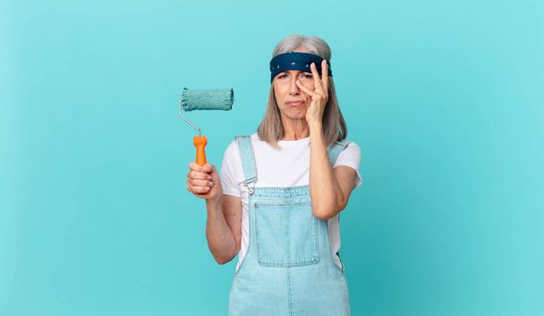 middle age white hair woman feeling bored, frustrated and sleepy after a tiresome with a roller painting a wall - 写真・画像