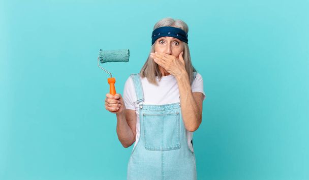 middle age white hair woman covering mouth with hands with a shocked with a roller painting a wall - Fotoğraf, Görsel