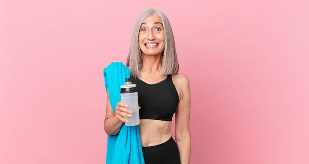 középkorú fehér hajú nő néz boldog és kellemesen meglepett egy törülköző és vizes palack. alkalmassági elv - Fotó, kép