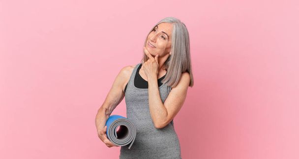 middle age white hair woman smiling with a happy, confident expression with hand on chin and holding a yoga mat. fitness concept - Foto, Imagem