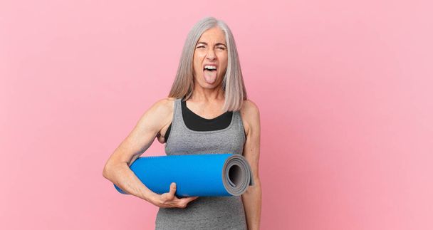 middle age white hair woman with cheerful and rebellious attitude, joking and sticking tongue out and holding a yoga mat. fitness concept - Foto, imagen