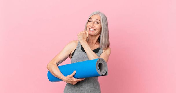 middle age white hair woman feeling happy and facing a challenge or celebrating and holding a yoga mat. fitness concept - Foto, imagen
