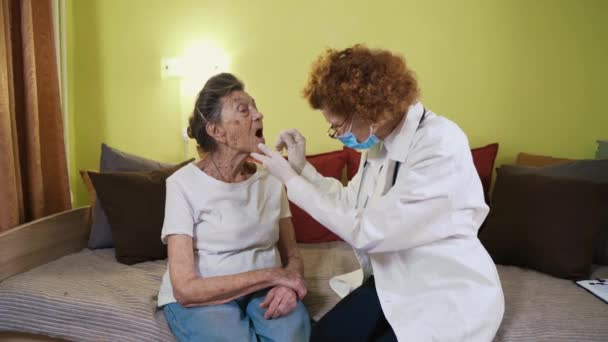 Medical professional in mask injects throat swab into sick elderly patient at home. Rapid antigen test analyze sample Covid-19, coronavirus pandemic at retirement home. Senior woman in quarantine - Filmati, video
