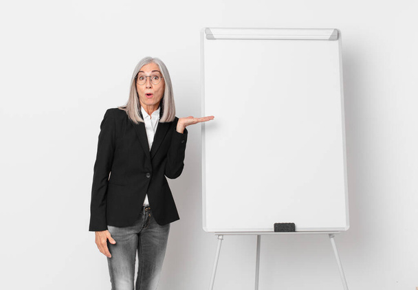 middle age white hair woman looking surprised and shocked, with jaw dropped holding an object and a board copy space. business concept - Zdjęcie, obraz