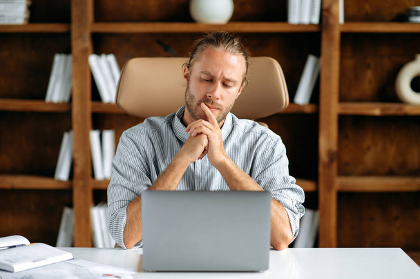 Pensive skupiony poważny młody dorosły mężczyzna kaukaski, dyrektor generalny lub pracownik biurowy, siedzi w biurze przy stole, za pomocą laptopa, spoczywa głowę na rękach myśli i analizy strategii biznesowej - Zdjęcie, obraz
