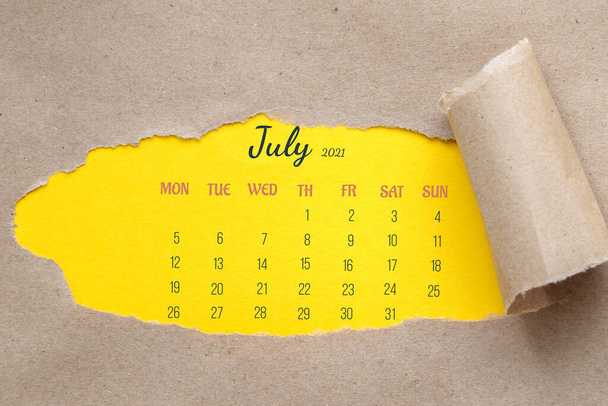 July 2021. Hole in paper with edges torn off. Through torn hole, you can see yellow background with calendar page for entire month. - Fotoğraf, Görsel