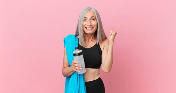 middle age white hair woman feeling shocked,laughing and celebrating success with a towel and water bottle. fitness concept - Photo, Image