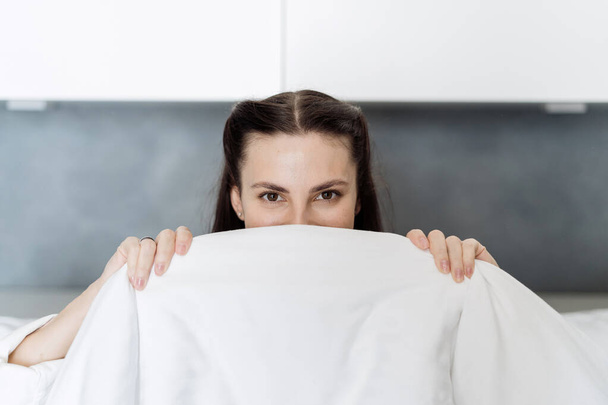Portrait of happy young woman cover her smiling face with white blanket, hides under soft duvet, spending good morning in cozy bedroom at home, looking at camera. Female rest and having fun in bed - Photo, image