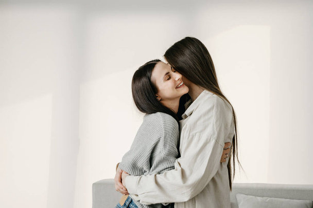 Side profile view of young girlfriends happy to meet each other, hugging, smiling wide, standing at home, against copy space background. Two cheerful woman cuddle, having fun and enjoying moment - Zdjęcie, obraz