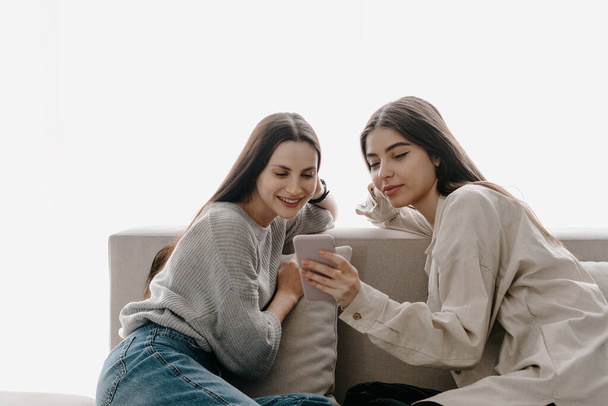 Две юные подруги сидят на диване, используя интернет-приложение на современном смартфоне, смотрят на дисплей и делают онлайн-покупки вместе. Концепция беспроводных технологий и социальных сетей - Фото, изображение