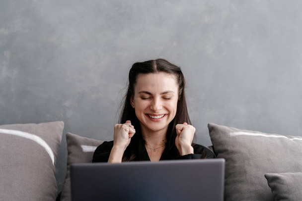 Portret młodej kobiety siedzącej z laptopem na wygodnej kanapie w salonie, podnoszącej pięści, radującej się i szeroko uśmiechniętej. Szczęśliwa kobieta świętuje zwycięstwo, robi ekscytującą minę - Zdjęcie, obraz
