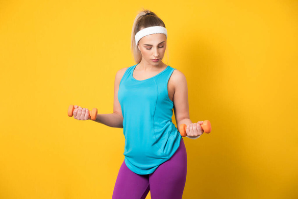 Sporty girl training biceps muscle with dumbbell, isolated on yellow background. Half length of fit sportswoman lifting weights. Fitness motivation. 80s styling. Studio shot - Φωτογραφία, εικόνα