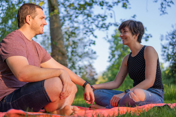 Photo d'un jeune et séduisant couple assis sur une serviette dans le parc se souriant et parlant. Grande journée d'été, attitude détendue. - Photo, image