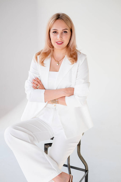 Confident young beautiful manager on white background. blonde girl in a white suit sits on a chair, on a white background. Strong and independent businesswoman. beautiful girl with a strong look. stylish girl in a trouser suit - Photo, image