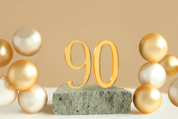Νούμερο 90 σε ένα βάθρο από μπετόν και ογκομετρικές χρυσές μπάλες σε μπεζ φόντο. Αντιγραφή χώρου - Φωτογραφία, εικόνα