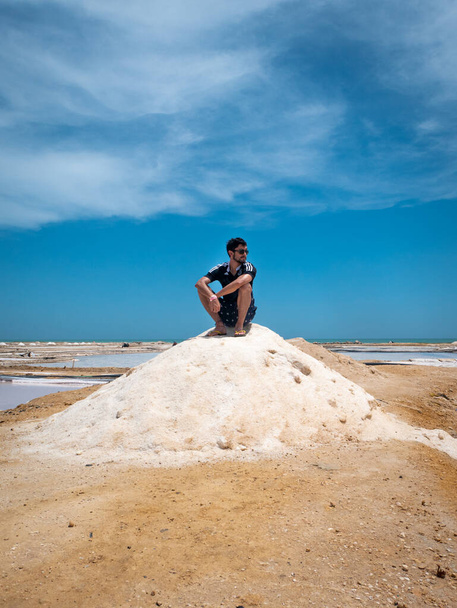 Manaure, La Guajira, Colombia - 29 de mayo de 2021: Joven latino sentado en una montaña de sal en las salinas marítimas más importantes del país - Foto, imagen