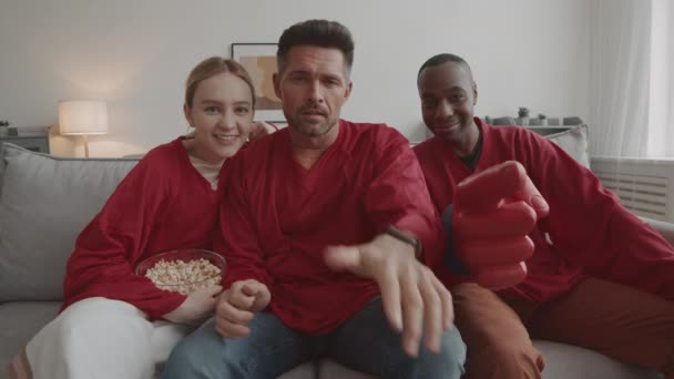 Medium long POV of diverse football fans wearing matching red long-sleeved jerseys, sitting on sofa at home, African man pointing foam finger on camera - Footage, Video