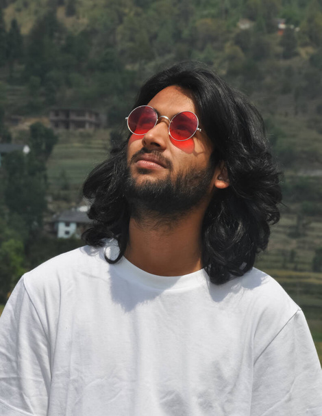Retrato de un guapo joven indio de pelo largo con gafas de sol mirando hacia arriba, un guapo barbudo parado afuera con una camiseta blanca  - Foto, imagen