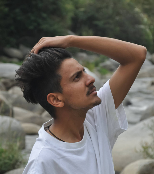 Un joven indio guapo parado al lado del río con una camiseta blanca pasando su mano por el pelo - Foto, Imagen