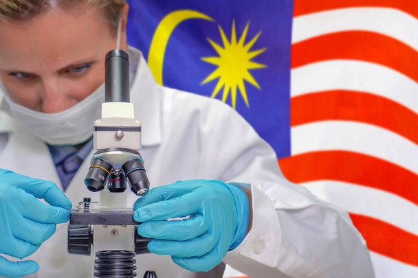 Biochimiste féminine regardant à travers un microscope sur fond de drapeau de Malaisie. Technologie médicale et recherche pharmaceutique et développement de concepts scientifiques en Malaisie - Photo, image