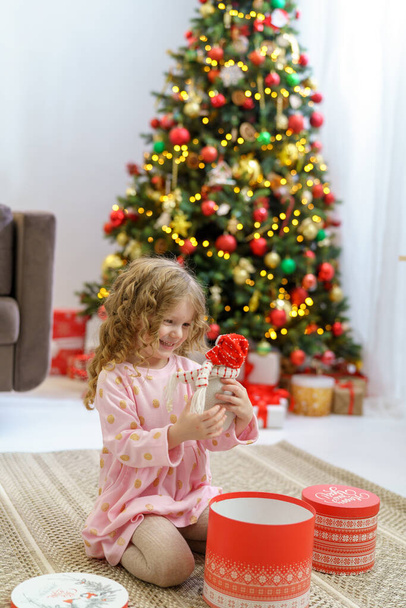 happy smiling girl with wavy hair holding a Christmas present near a charming Christmas tree. the child is dressed in a dress against the background of a garlanded house and Christmas lights. top view - 写真・画像