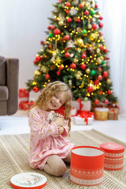 happy smiling girl with wavy hair holding a Christmas present near a charming Christmas tree. the child is dressed in a dress against the background of a garlanded house and Christmas lights. top view - Photo, image