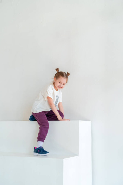 A little happy girl in fashionable clothes smiles and poses against the white wall of the studio. concept of advertising children's clothing, emotional children, stylish children's image. Copy space - Photo, image