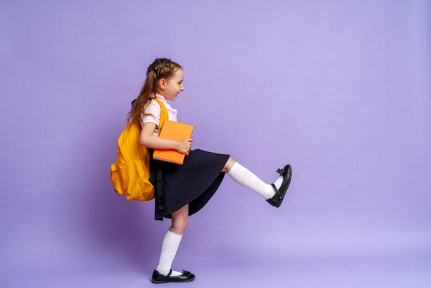 Cute happy kid in uniform. raises his leg high and runs on purple background. child with backpack. little girl is ready for school. Dynamic images that go back to conceptual school. holidays begin. - Photo, image