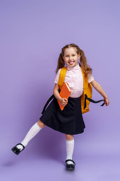 Cute happy kid in uniform. raises his leg high and runs on purple background. child with backpack. little girl is ready for school. Dynamic images that go back to conceptual school. holidays begin. - Zdjęcie, obraz