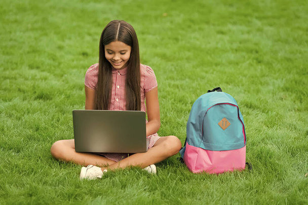 happy kid using laptop for online school education sitting in park on green grass with backpack, childhood - Foto, Bild