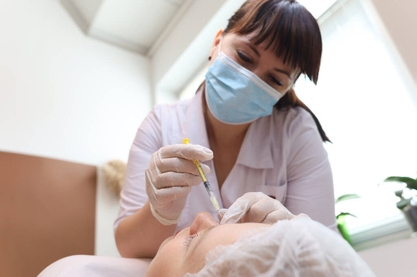 The cosmetologist leaning over the client performs a chin lift procedure by injection cosmetic injections - 写真・画像