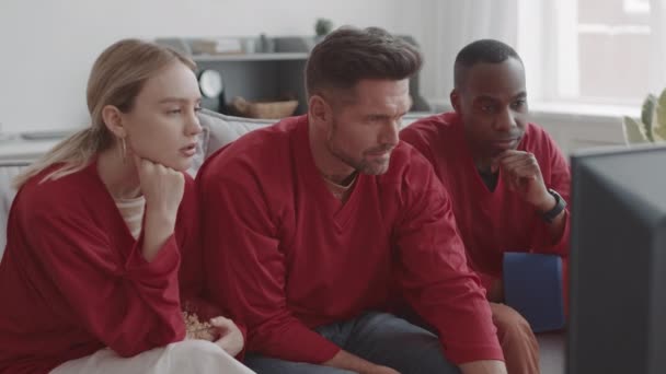 Medio lungo di tre diversi appassionati di sport che indossano magliette rosse a maniche lunghe, seduti sul divano a casa, guardando la televisione tranquillamente - Filmati, video