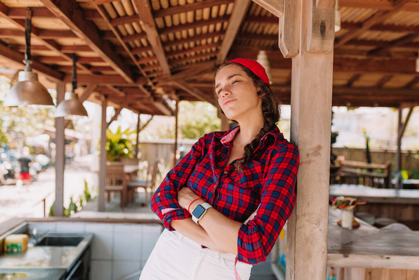 Elegante donna hipster con cappuccio rosso e camicia tesa ha piegato le mani di fronte a lei e posa sulla terrazza estiva in legno - Foto, immagini