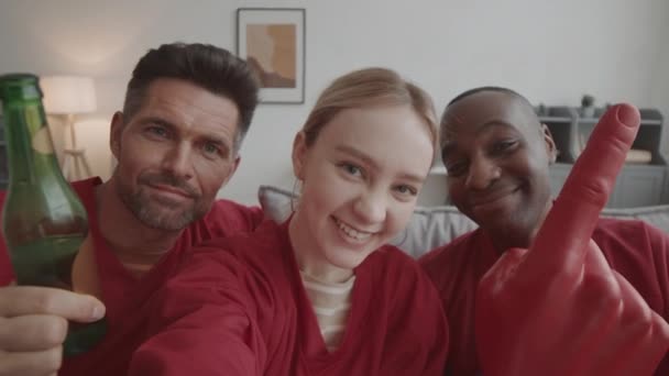 Chest-up handheld POV of blond-haired happy woman wearing matching red long-sleeved jerseys with her male Caucasian and African friends, recording selfie video at home - Footage, Video