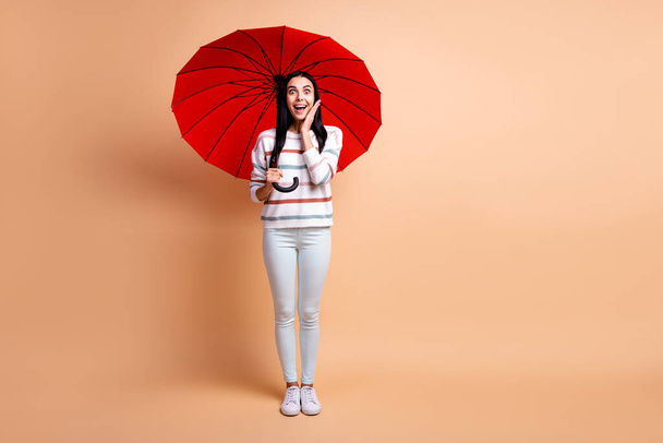 Full size photo of young happy positive smiling girl with big red parasol hold hand on cheek isolated on beige color background - Foto, afbeelding